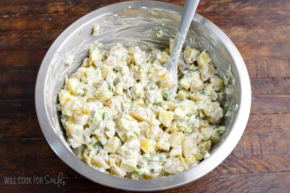 Dill pickle potato salad tossed in creamy dressing in a mixing bowl with a spatula.