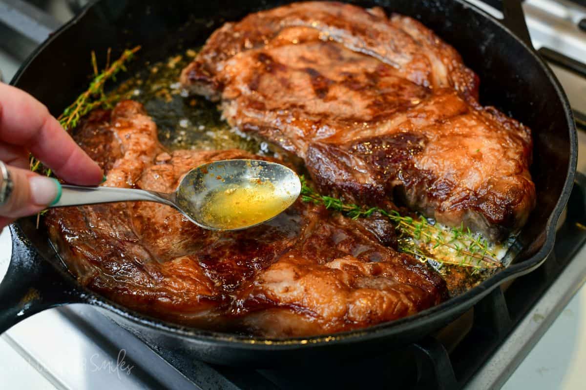 Hand basting seared ribeye steaks with melted butter and thyme in a cast-iron skillet for added flavor and crust.