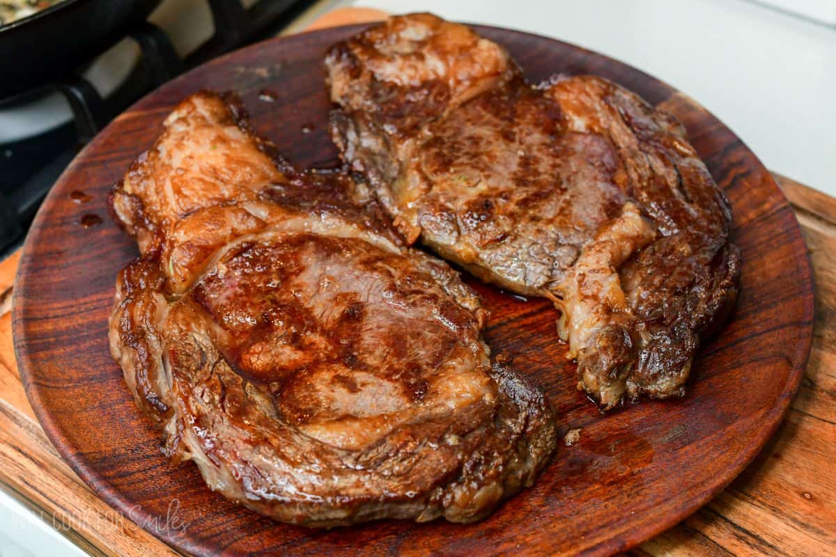 Dry brined ribeye steaks resting on a wooden platter after cooking, allowing juices to redistribute before slicing and serving.