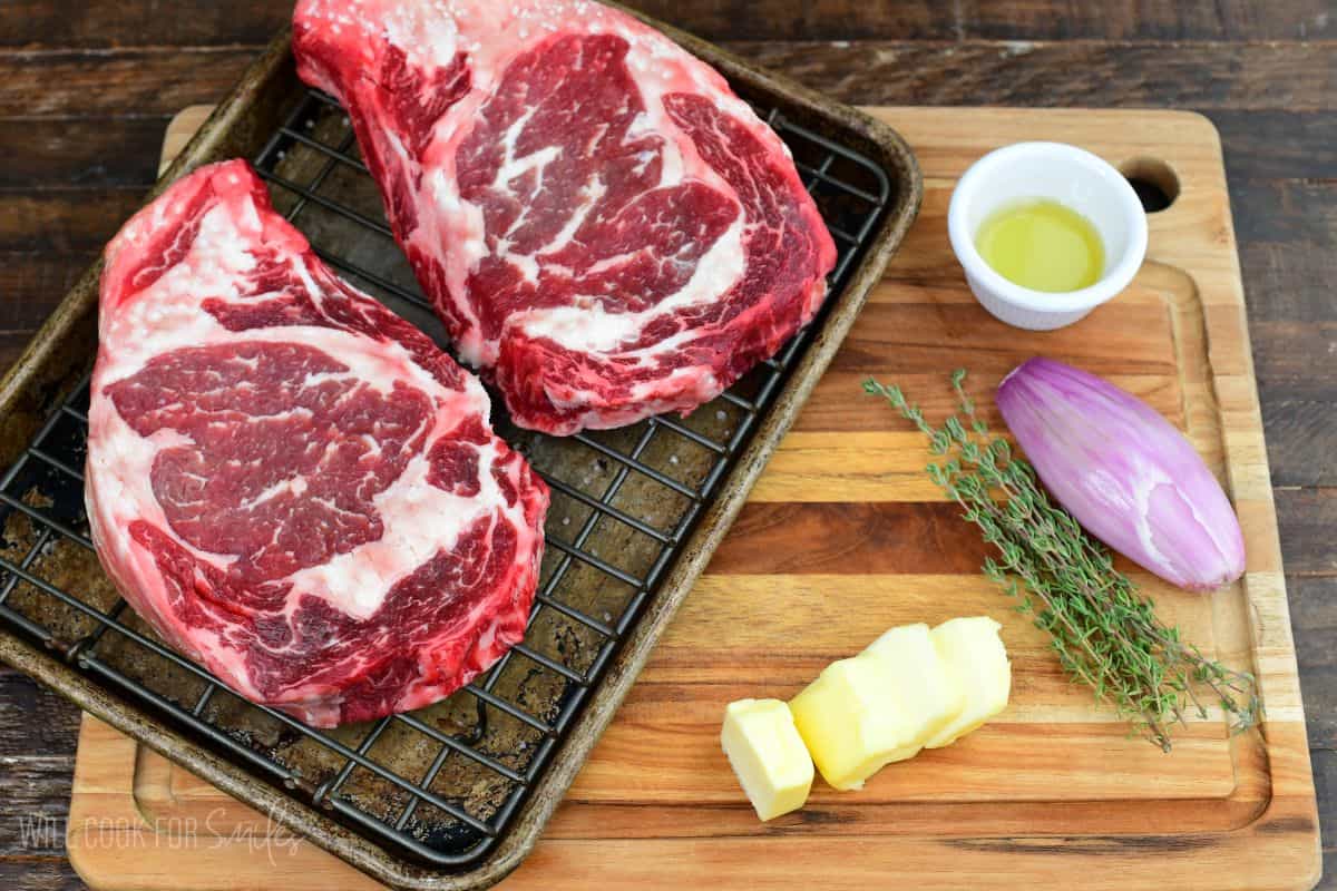 Ingredients for dry brined ribeye steak, including thick-cut ribeyes, butter, shallot, thyme, and oil arranged for cooking preparation.