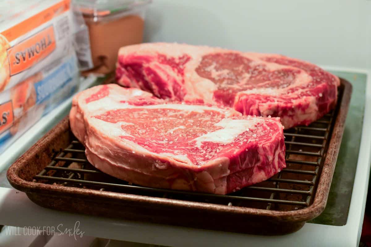 Two ribeye steaks dry brining on a wire rack in the refrigerator, seasoned with salt to improve flavor and tenderness.