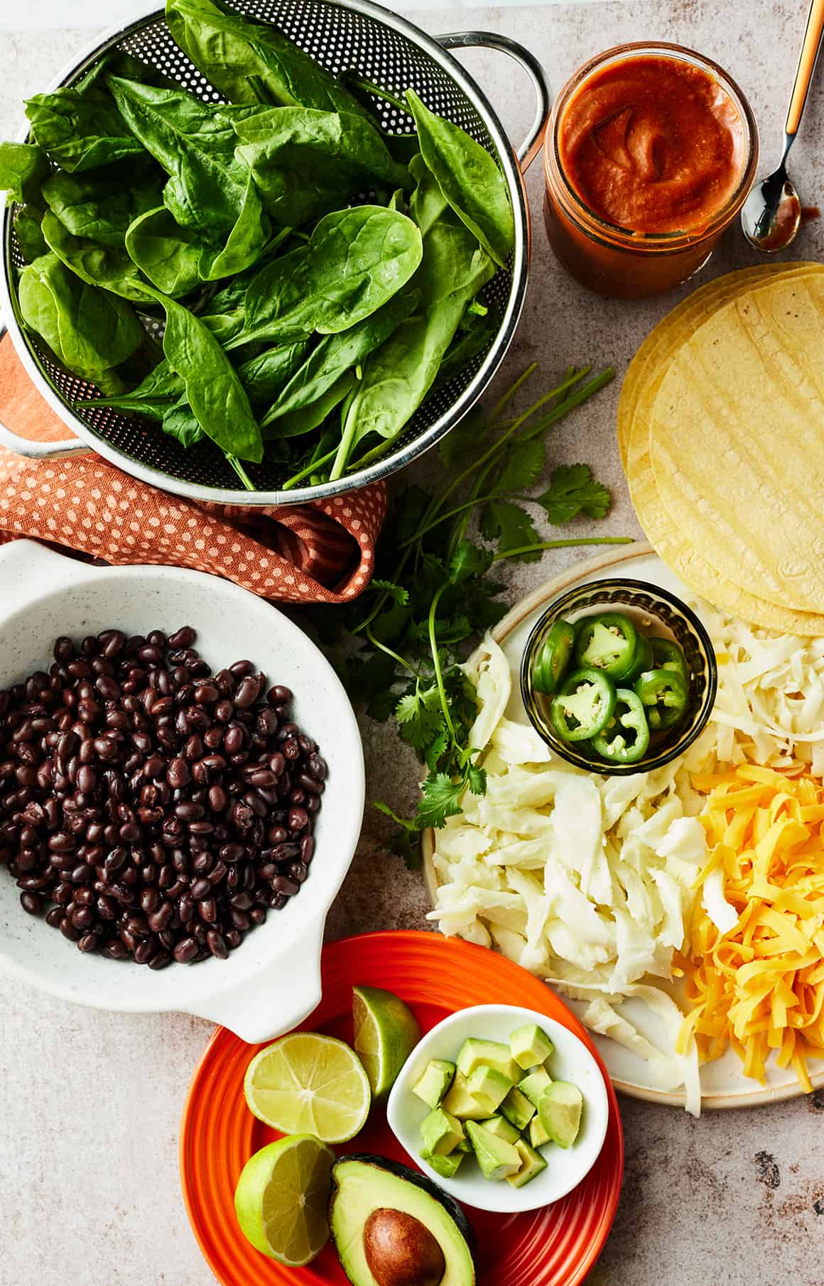Overhead shot of spinach in colander, bowl of black beans, jar of sauce, corn tortillas, plate of shredded cheese, avocado, and limes