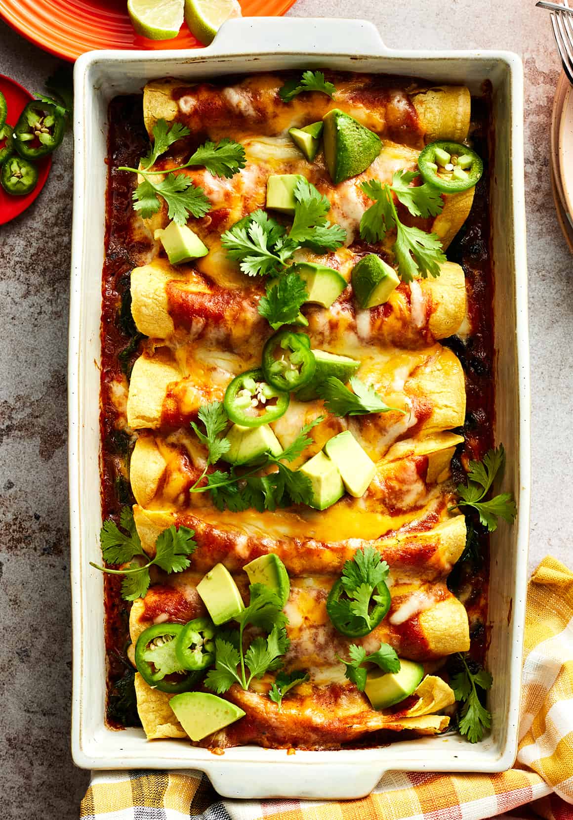 Easy enchiladas in baking dish topped with sauce, melted cheese, avocado, jalapeños, and cilantro