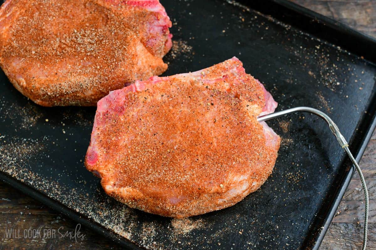 Seasoned bone-in pork chops on a baking sheet, with a thermometer probe inserted into one chop to monitor cooking temperature.