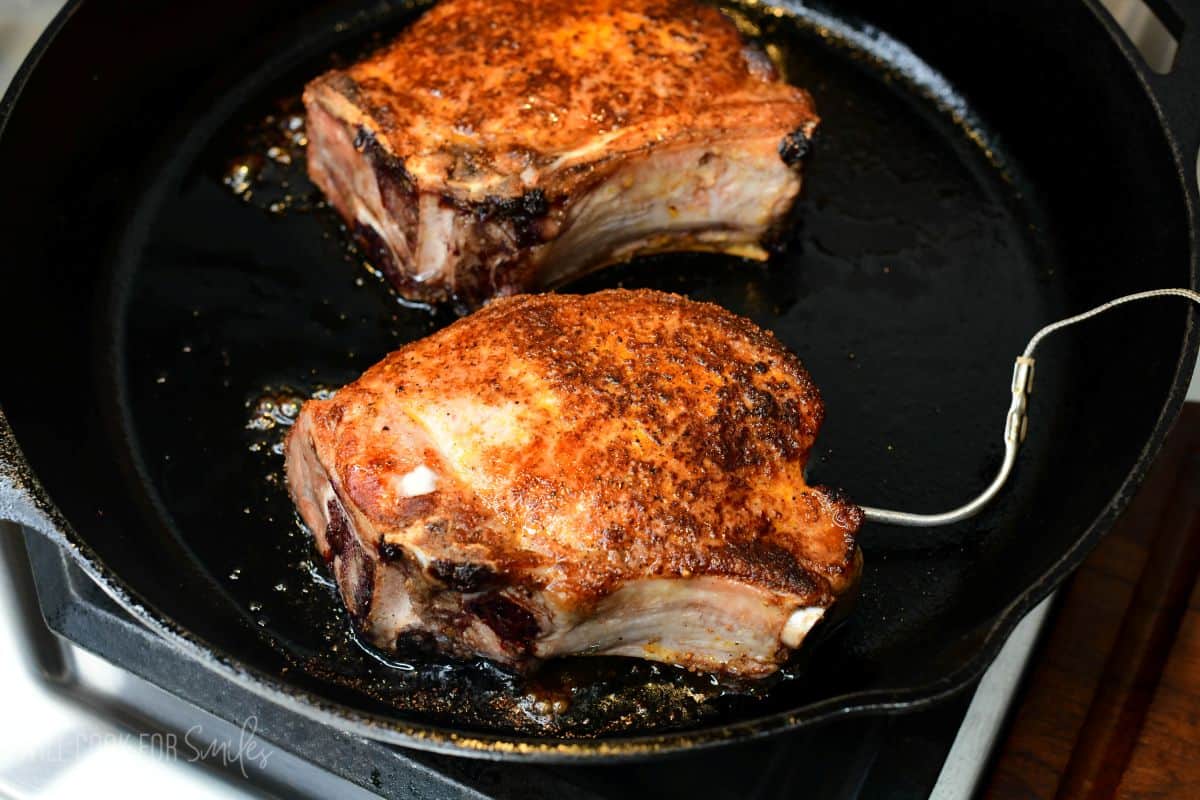 Bone-in pork chops coated in seasoning searing in a cast-iron skillet, with a thermometer probe monitoring temperature.