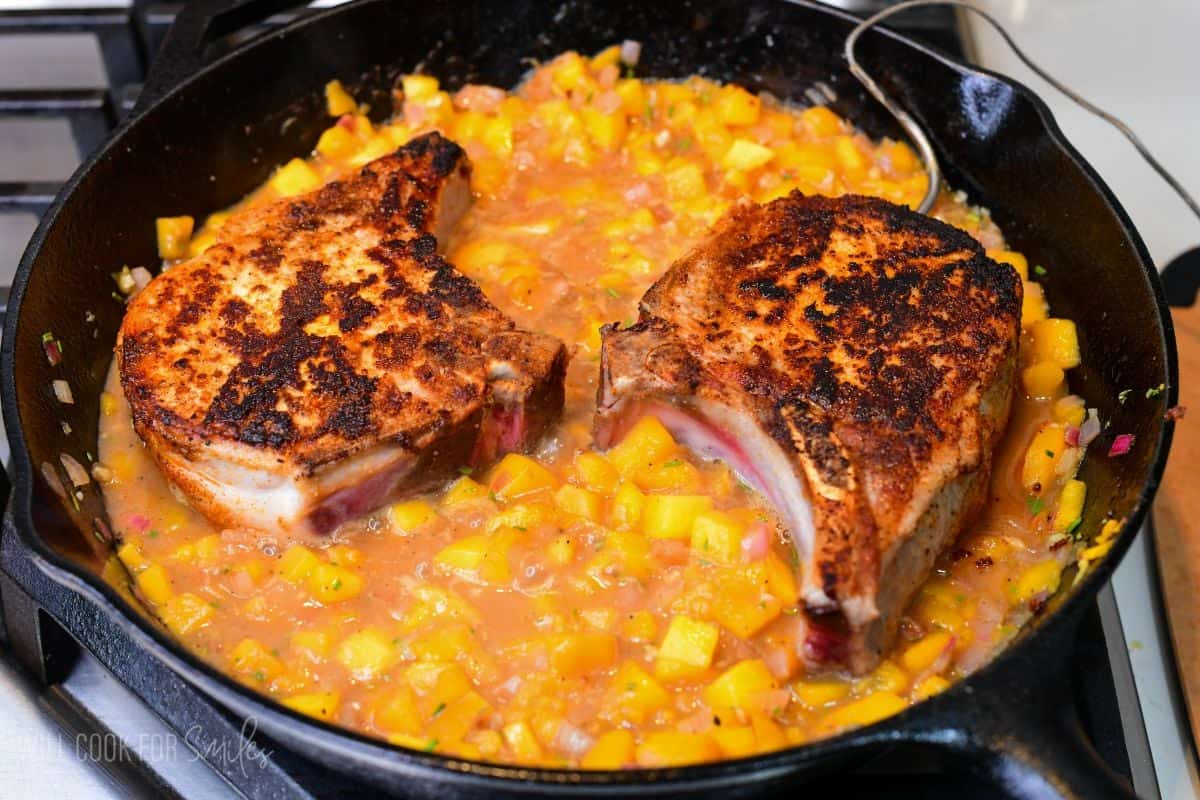 Pan-seared pork chops nestled in a peach sauce with diced peaches, simmering in a cast-iron skillet.