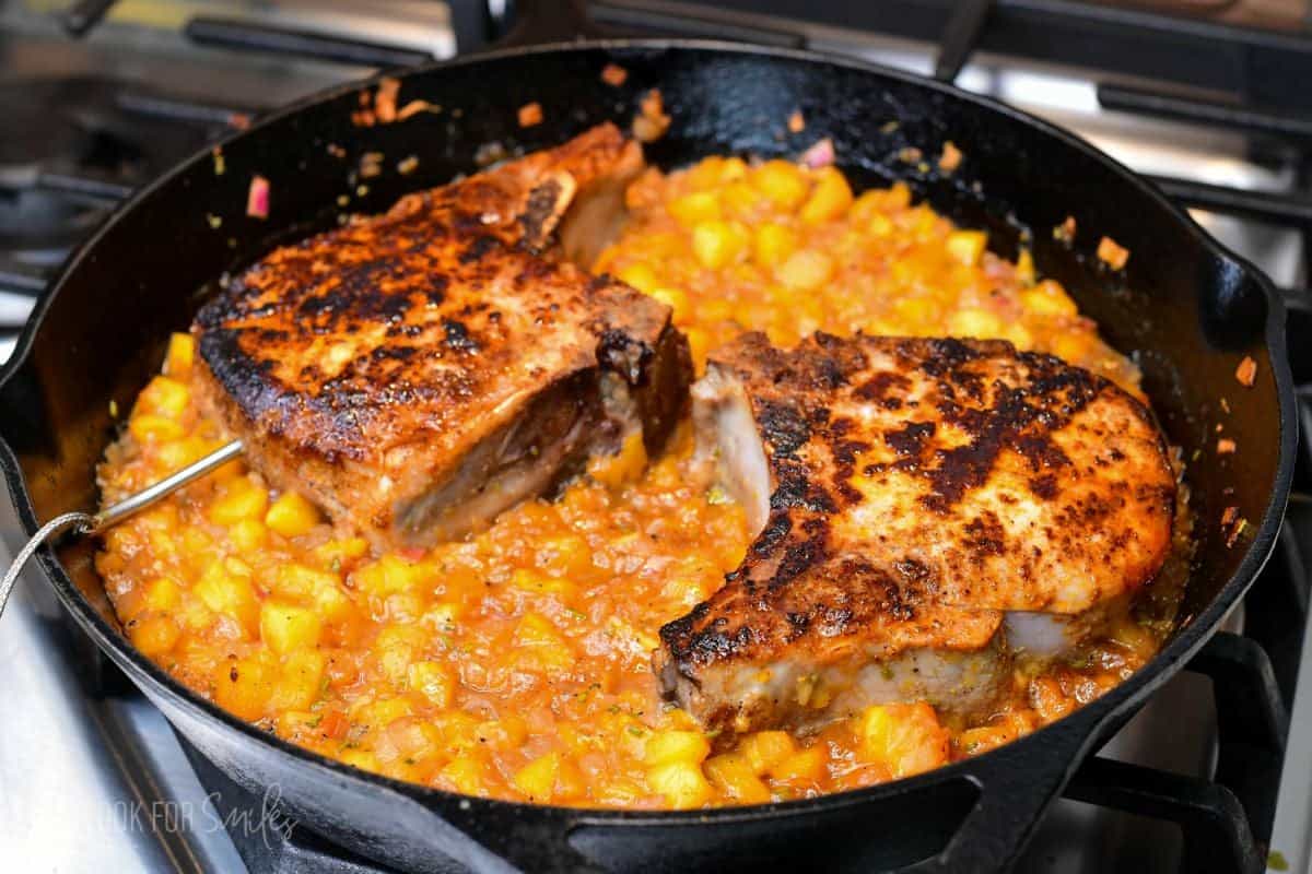 Peach pork chops finishing in the oven with a chunky peach sauce in a cast-iron skillet.