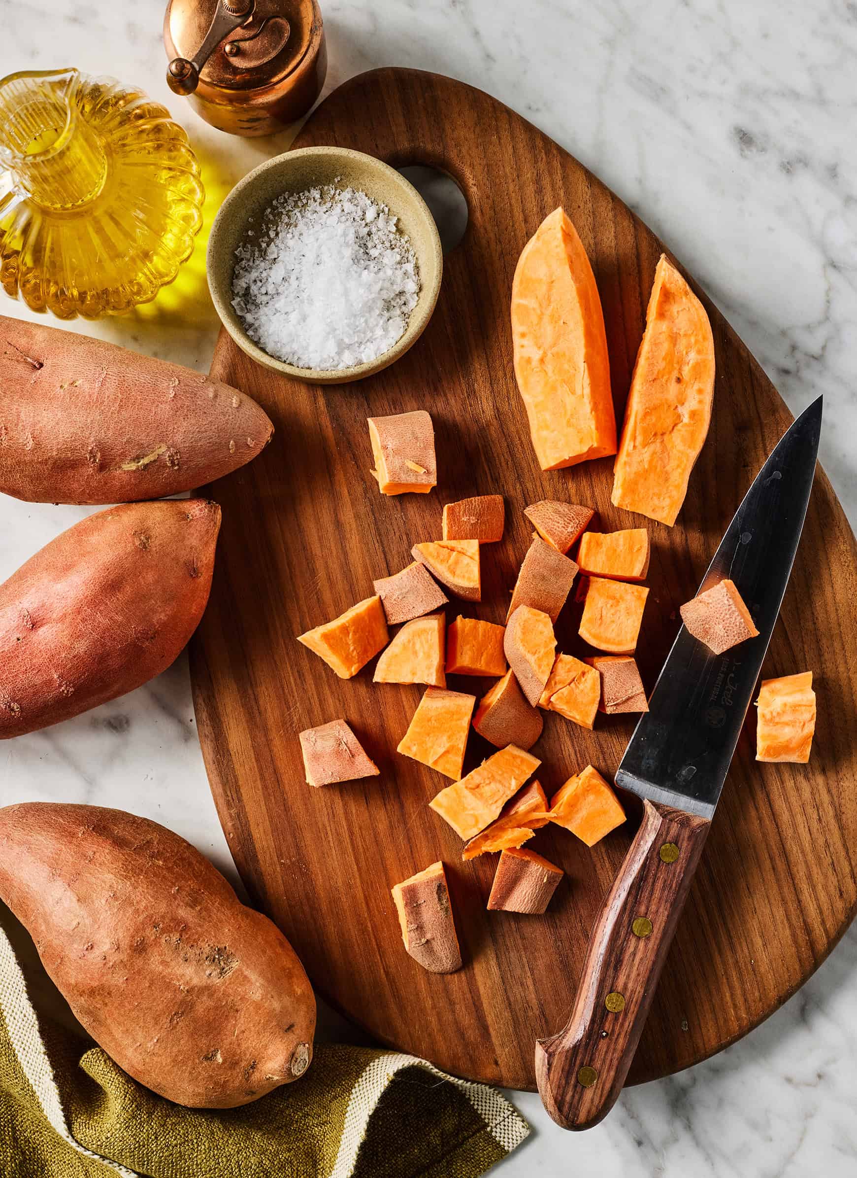 Chopped sweet potatoes on cutting board next to olive oil, salt, and pepper grinder