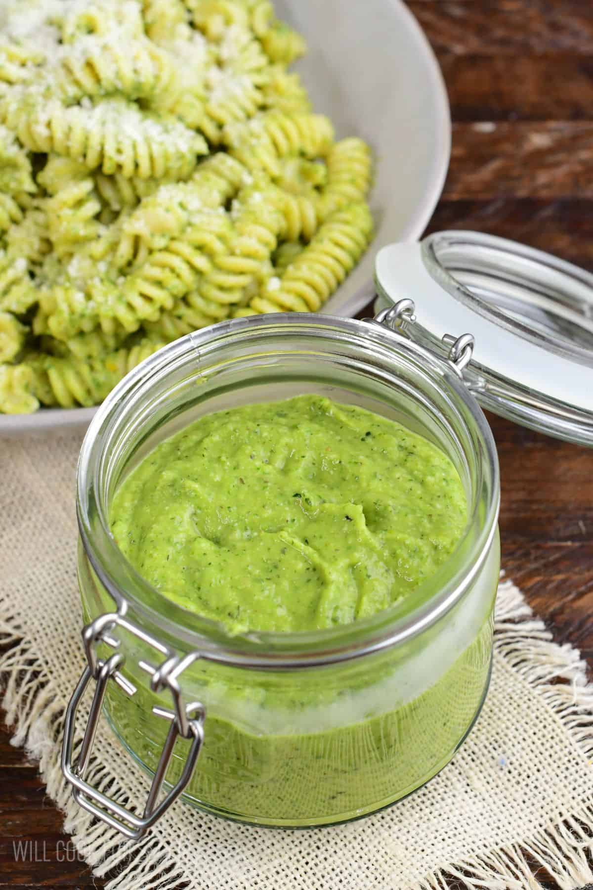 Creamy homemade zucchini pesto in a glass jar with zucchini pesto pasta in the background.
